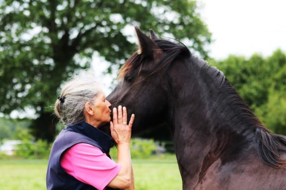 Femme blonde embrassant un grand cheval noir dans un environnement naturel.